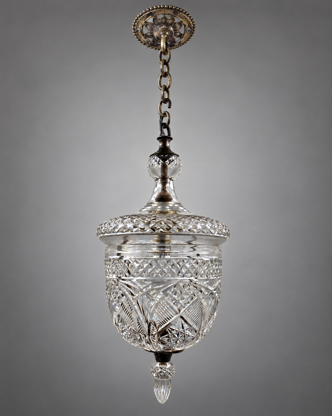 An antique cut glass pendant light with ornate silvered metal hardware, hanging against a neutral gray background.